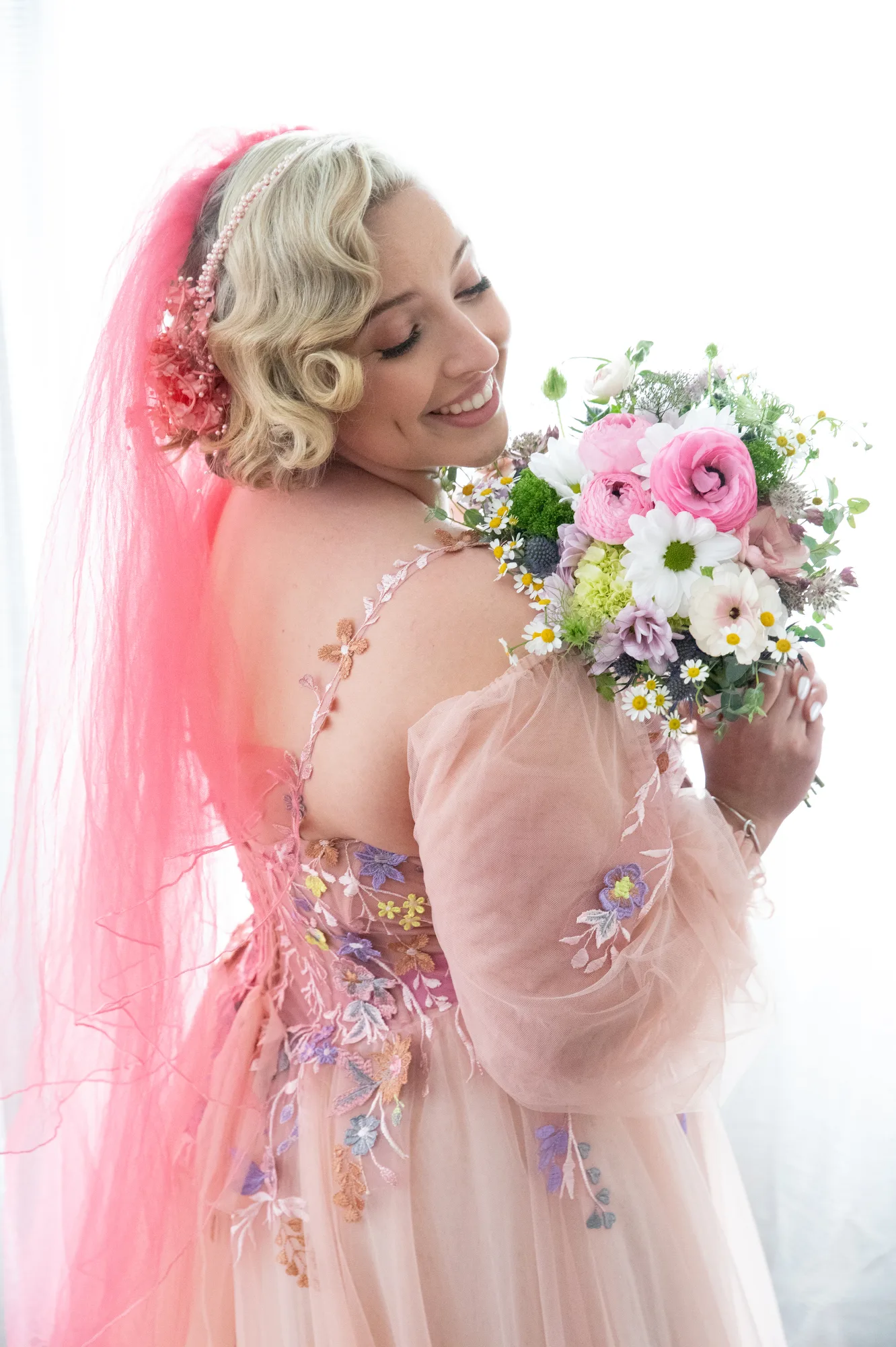 A bride wearing a pink dress and pink veil holds her bouquet and smiles at the Swan Club on Long Island.