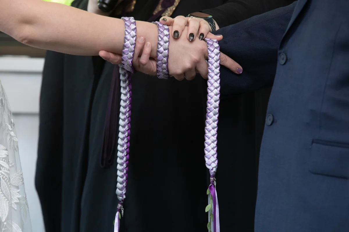 A non-binary couple holds hands wrapped in a hand fasting cord at their New York wedding.