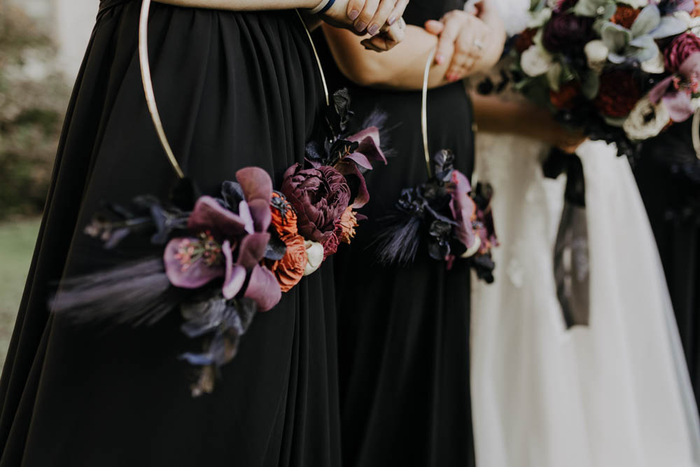 Bridesmaids in black dresses holding gold hoops with burgundy, orange, and purple flowers.