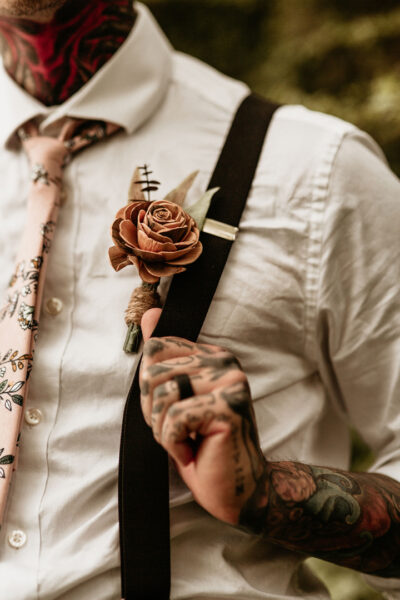 Close up of groom's tattooed arm and hand on a black suspender with a tan wood rose boutonniere.