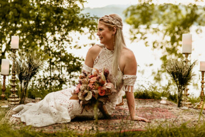 Bride sitting on a rug in the woods, holding a bouquet of wood flowers. The area is decorated with gold candlesticks, for an elopement.