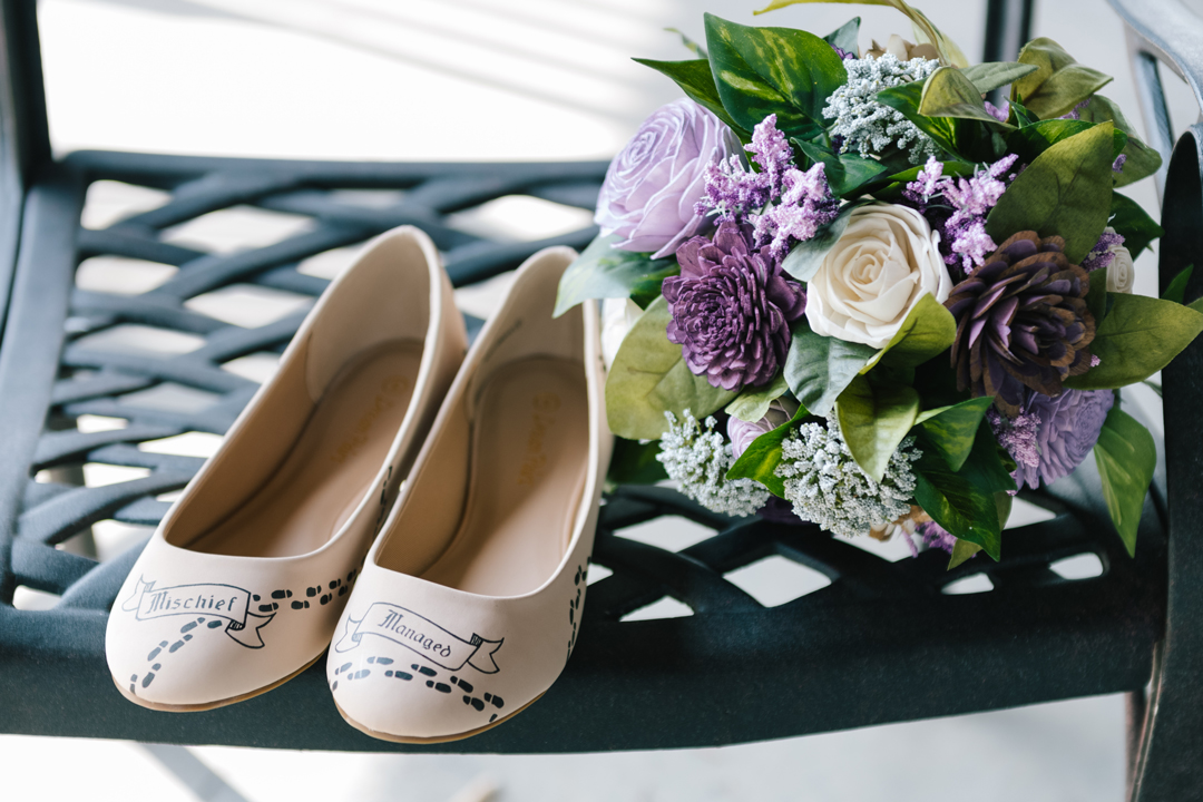 Bride's shoes that say "Mischief Managed" resting next to a bouquet of wood flowers dyed various shades of purple and ivory.