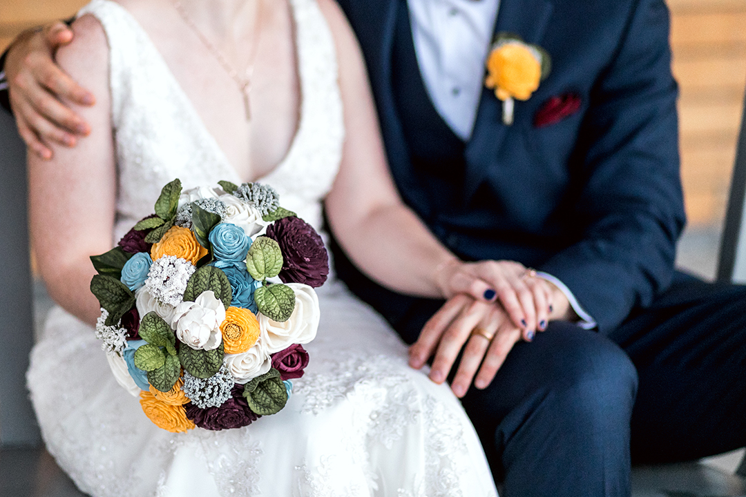 Bride and groom, sitting together outside. She's holding a bouquet and he has his arm around her.