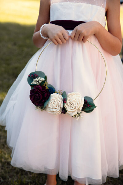 Flower girl in white dress holds a gold hoop with ivory and burgundy wood flowers.
