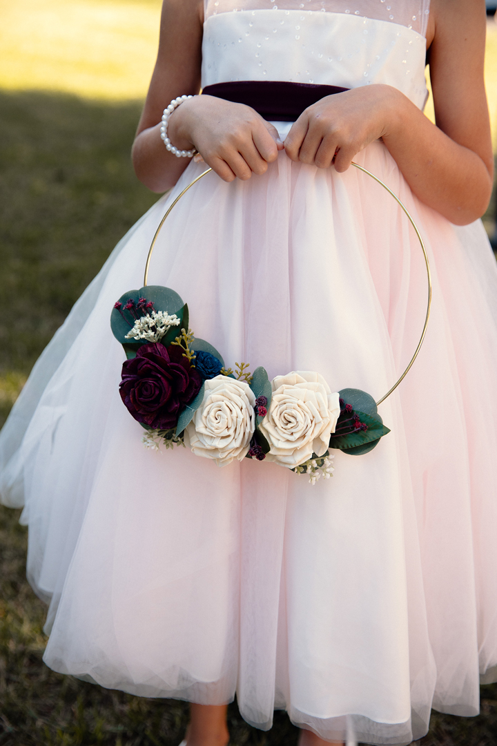 Flower girl in white dress holds a gold hoop with ivory and burgundy wood flowers.