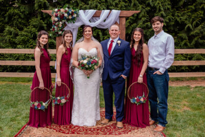 Couple and wedding party, standing in front of their ceremony arch. Bride is holding a bouquet while bridesmaids hold wreaths with wood flowers.