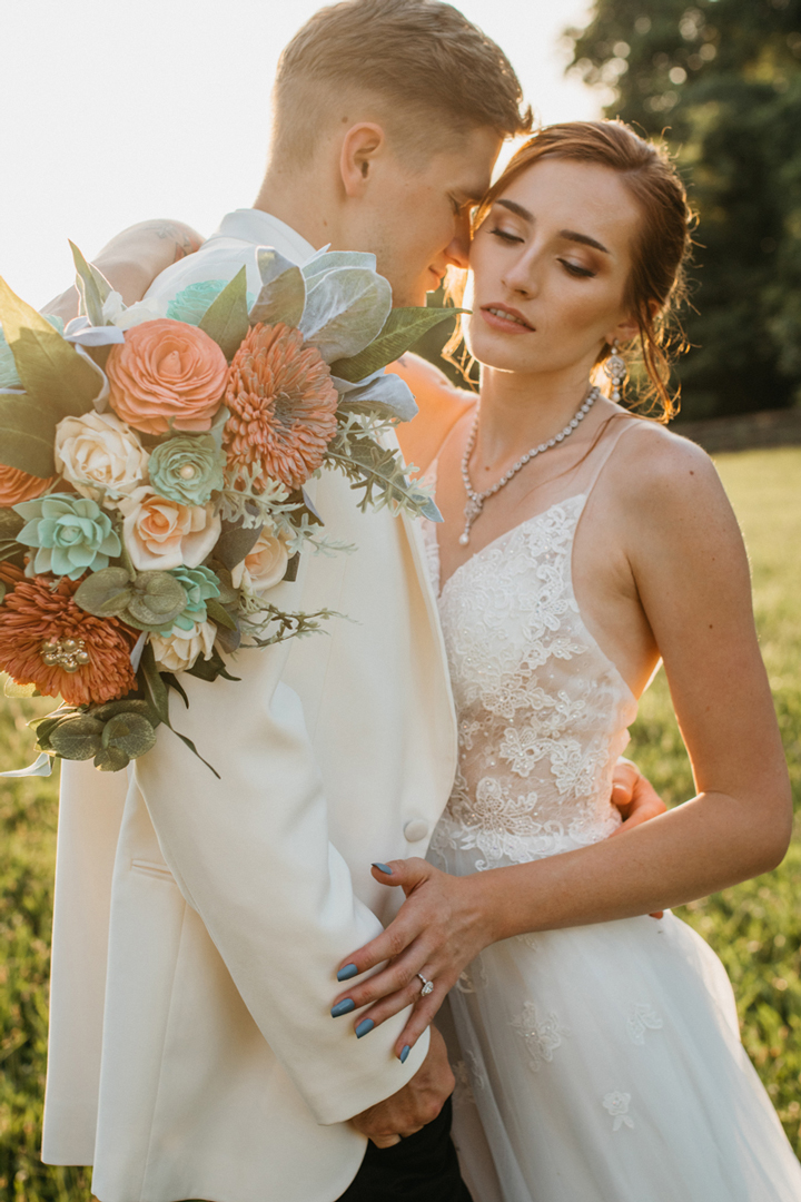 Bride and groom embracing outside. She is holding a bouquet of ivory, coral pink, and seafoam blue wood flowers.