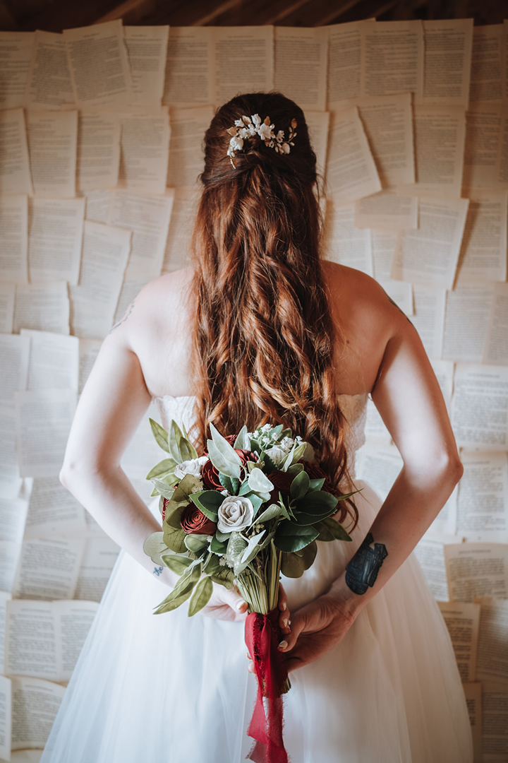 Tattooed bride with long red hair, holding a bouquet of red roses & book page flowers behind her back.