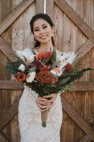 Smiling bride holding a bouquet of ivory, rust, and terracotta wood flowers out, toward the camera.