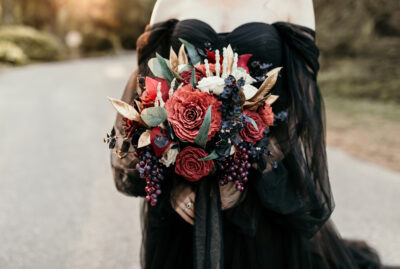 Bride in black dress, holding a large spooky bouquet of wood flowers including a faux skeleton hand and gold accents.