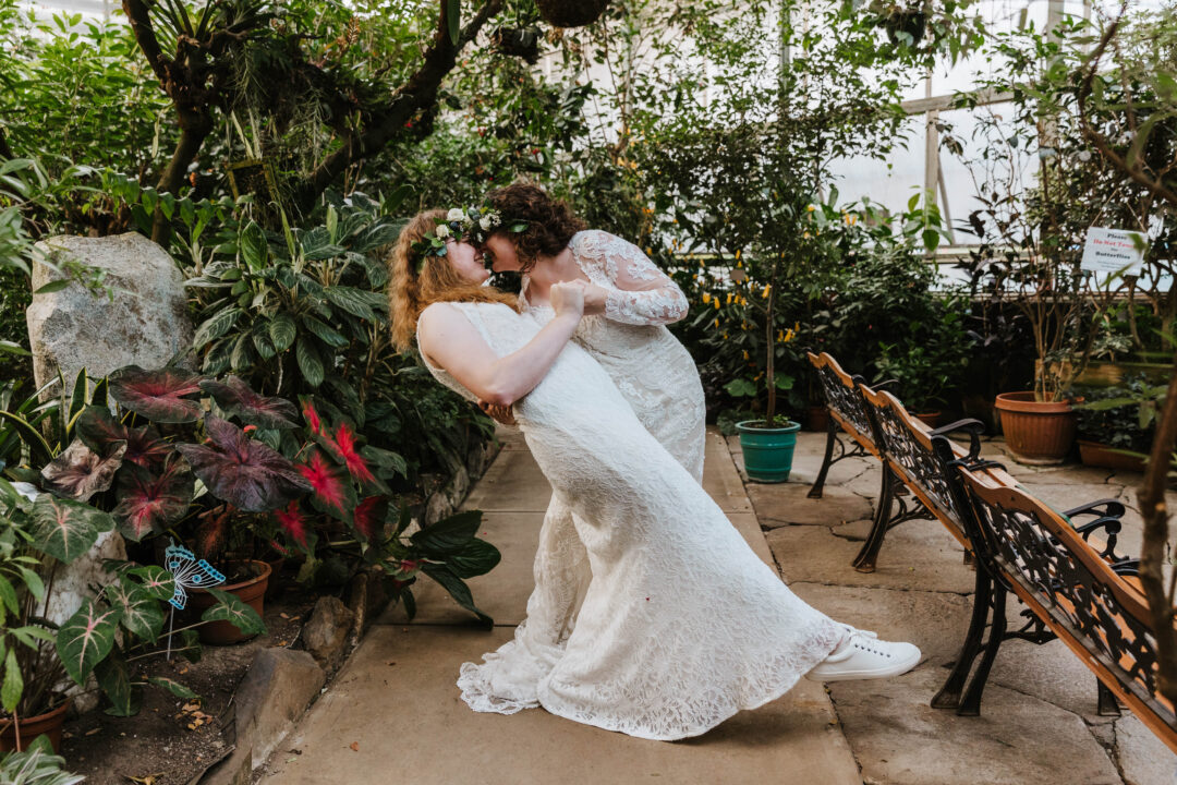 two just married brides do a dip while standing inside of a butterfly conservatory. they are both wearing white dresses and flower crowns