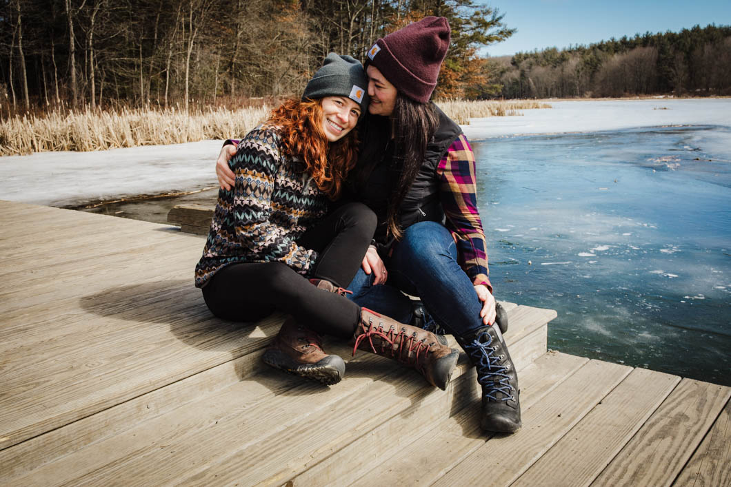 two lovers sit close to each other, snuggled up on a dock overlooking the lake behind them. There is snow & ice frozen over the water.