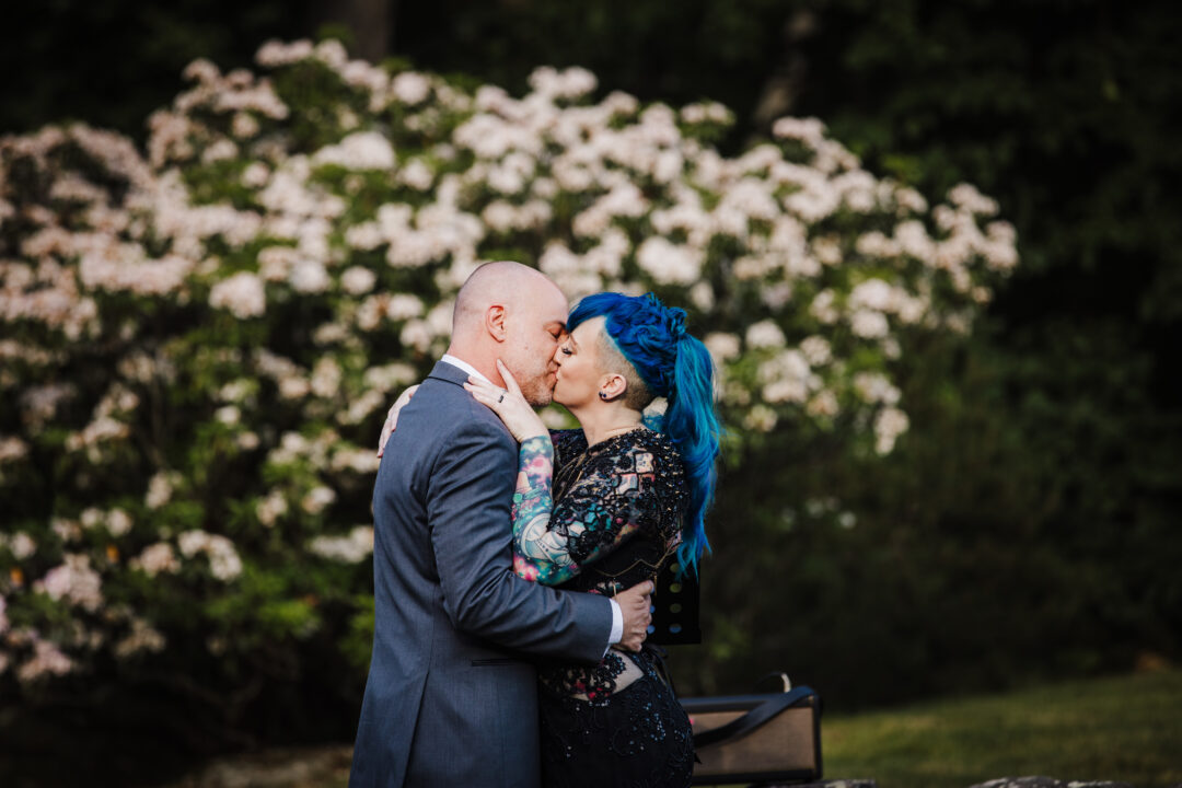 two newlyweds kiss in front of a white floral bush, one is wearing a suit and the other has on a black dress and blue hair