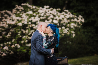 two newlyweds kiss in front of a white floral bush, one is wearing a suit and the other has on a black dress and blue hair