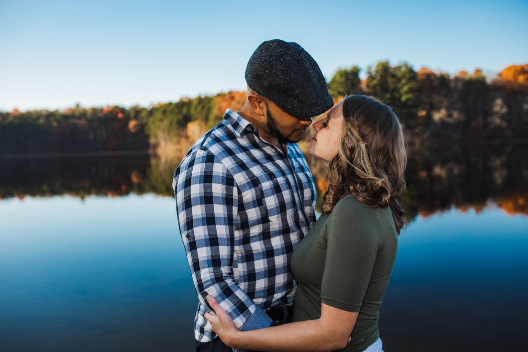 two lovers lean in for a kiss, they are standing in front of a body of water that has an autumn colored treeline behind the water.