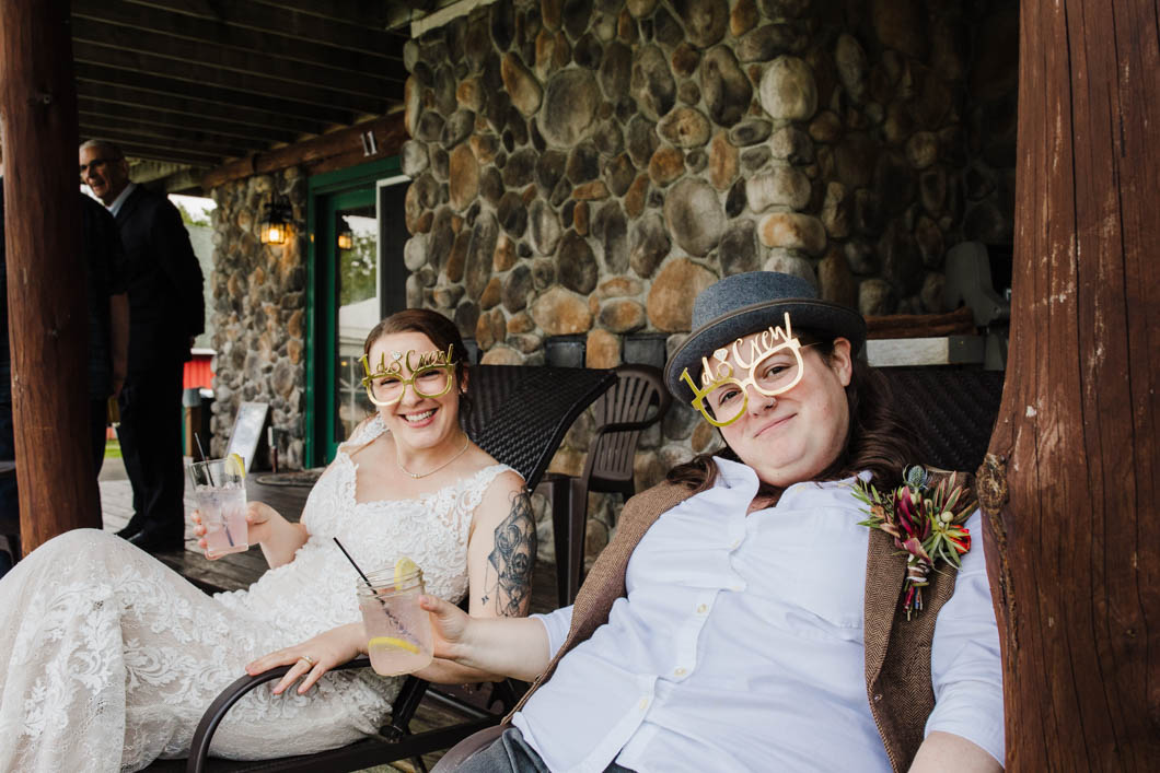 Two married people sit in chairs while enjoying a moment of relaxation during their wedding. They are both wearing party glasses that say "I do crew"