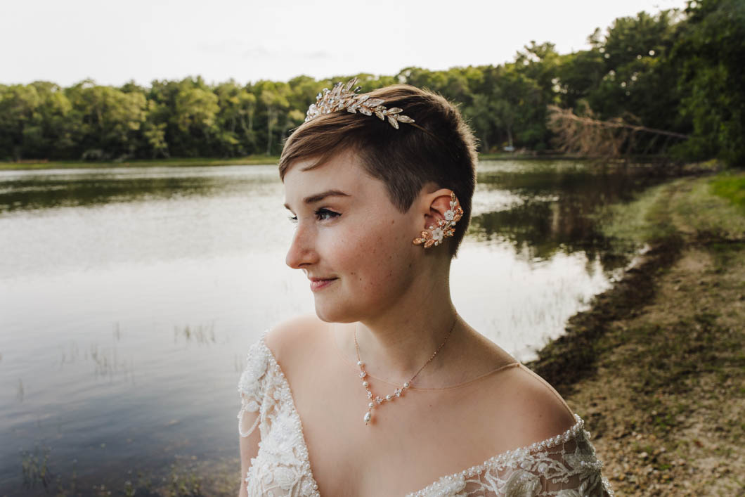 a person who just got married stands front of a body of water. they are wearing a white dress, golden tiara with gems stones on it as well as, an ear cuff with small white floral designs