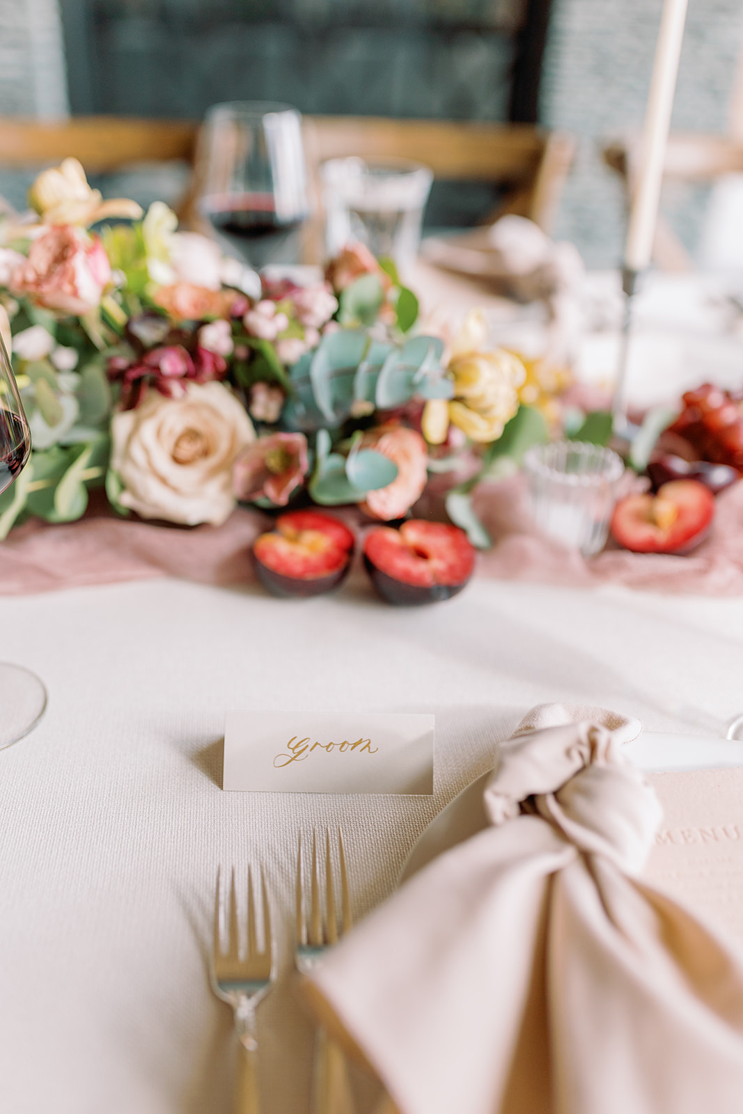 A wedding reception table, draped with a ivory textured linen, airy blush runner and a garland mixed with romantic florals, plums, pears and grapes.