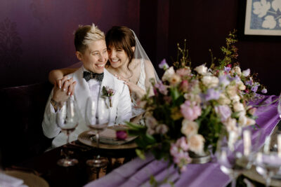 A couple in a white suit and white dress with a veil sit at a luxurious, intimate reception table adorned with a velvety purple runner, garden-inspired flowers and gold accents.