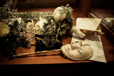 On a flat surface, the pillow for the rings, in black velvet with a gold embroidery; a white and gold mask and the peony bouquet behind.