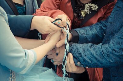 The hands of four people: the spouses holding together, the celebrants tying them with a black, blue and silver ribbon.