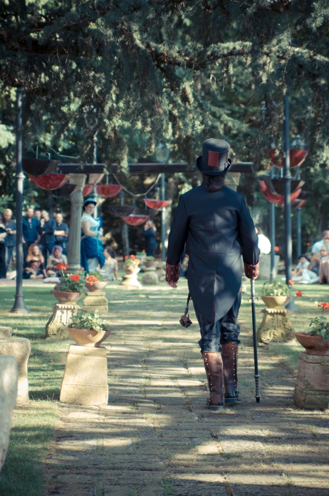 Black and burgundy steampunk groom surrounded by umbrellas hanged open and upside down, walking towards the ceremony, right in front of the guests.