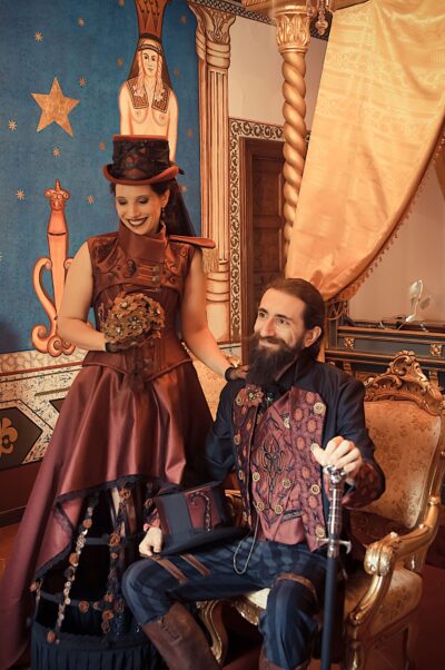 The bride stands, showing off her black and burgundy gown that is shorter in the front and shows the decorated cage that keeps the skirt wide, the groom sits down in his sepia and burgundy embroidered long tails suit.