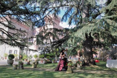 On the background, a pink rock castle and a lot of trees, in Tuscany. At the center, a steampunk bride walks towards the ceremony