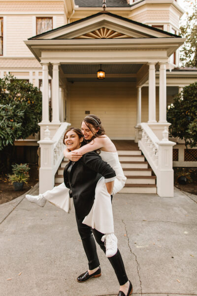 A bride in a black suit carries a bride in white in front of an ornate porch