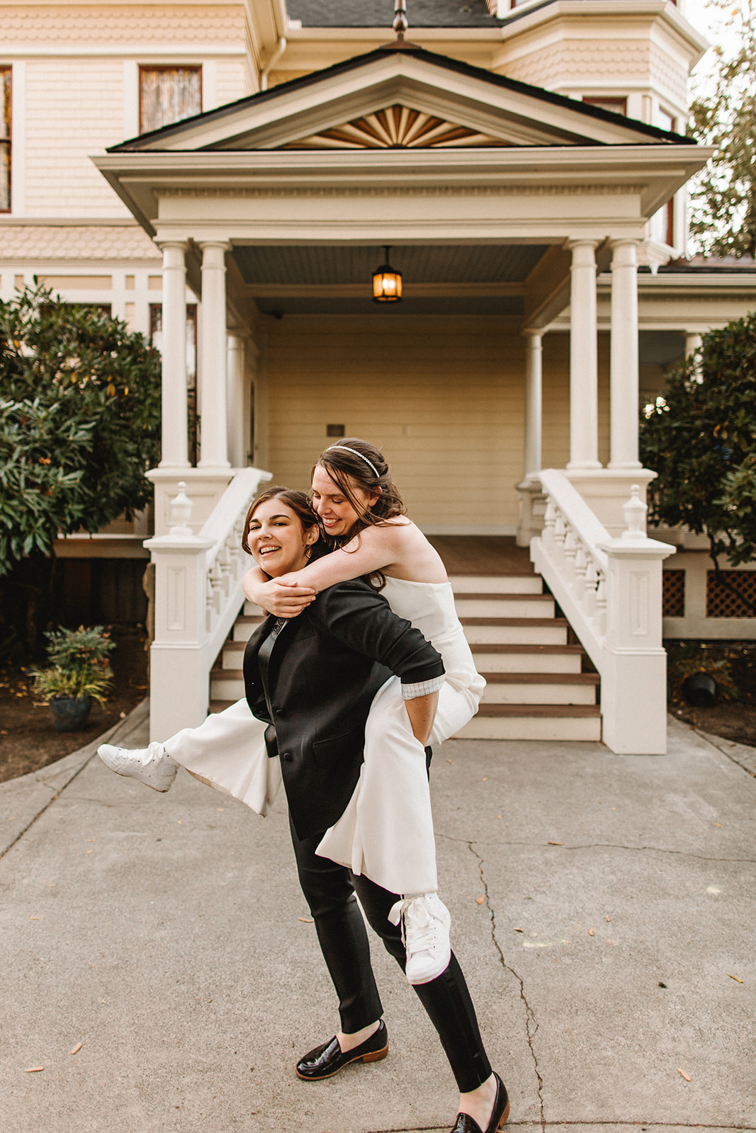 A bride in a black suit carries a bride in white in front of an ornate porch