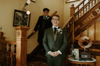 A first look photo on the grand staircase at the Victorian Belle Mansion in Portland, Oregon. A groom stands smiling as another groom descends the stairs behind him. Photo by Hannah Alena.