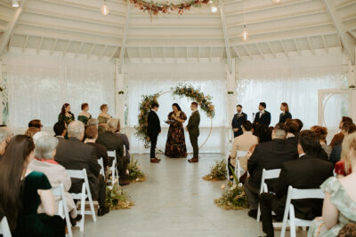 A wedding ceremony in the gazebo of the Victorian Belle Mansion in Portland, Oregon. Two grooms stand with their wedding party under a round arbor decorated with flowers and mushrooms. Photo by Hannah Alena.