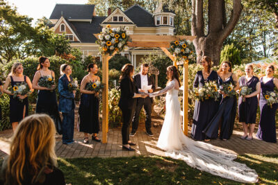 A wedding ceremony with one bride in a white dress with a long train and another bride in a black suit. The wedding party members surrounding them wear differently styled navy blue outfits and carry bouquets. A Victorian mansion stands in the background