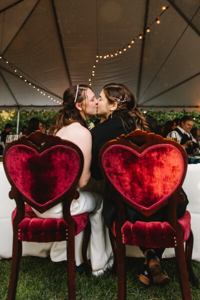 Two brides kiss while sitting in velvet-backed heart-shaped chairs