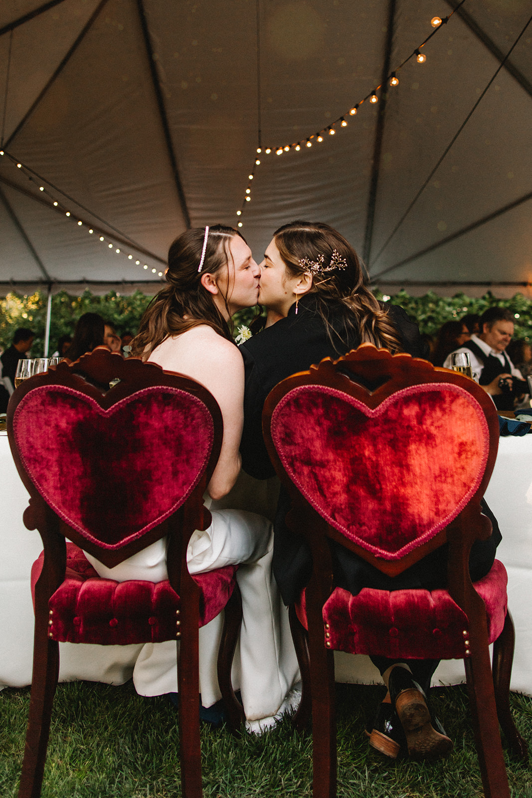 Two brides kiss while sitting in velvet-backed heart-shaped chairs