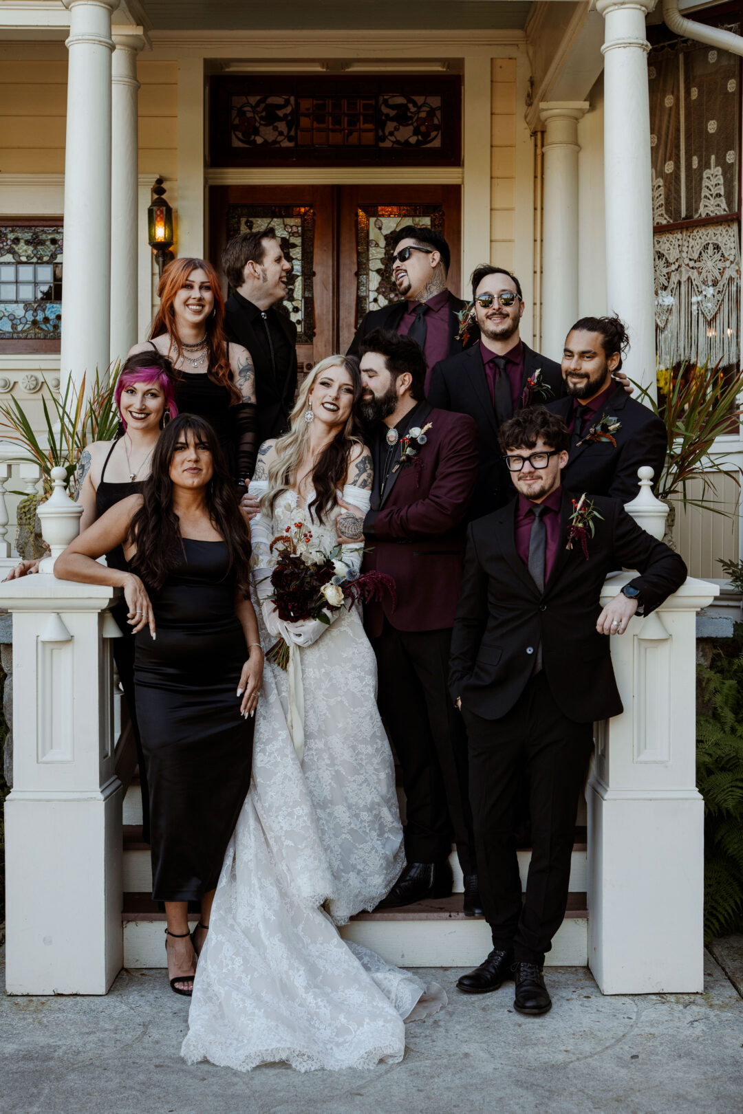 A wedding party stands on the stairs of the North Porch of the Victorian Belle Mansion in Portland, Oregon. The bride wears a white lace gown with gloves and the groom & wedding party wear black and burgundy.