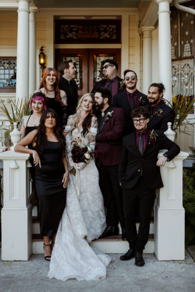 A wedding party stands on the stairs of the North Porch of the Victorian Belle Mansion in Portland, Oregon. The bride wears a white lace gown with gloves and the groom & wedding party wear black and burgundy.