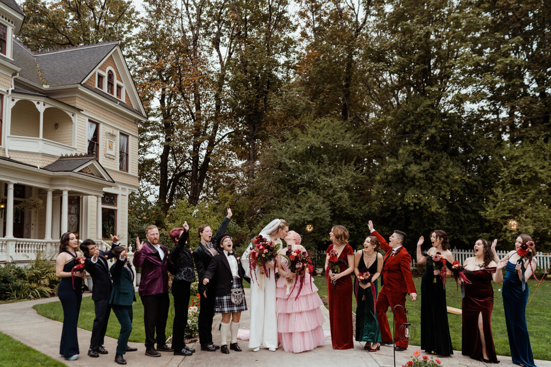 A wedding party stands in front of a Victorian mansion in Portland, Oregon. The couple kisses while the wedding party smiles.