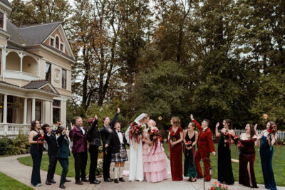 A wedding party stands in front of a Victorian mansion in Portland, Oregon. The couple kisses while the wedding party smiles.