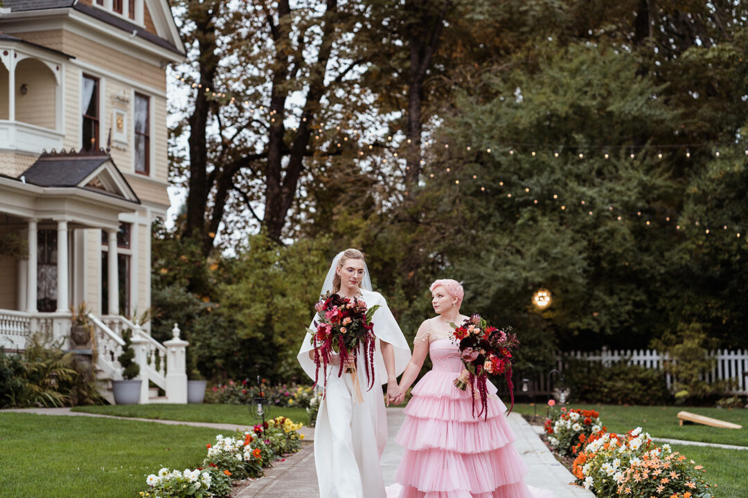 A couple walks into their ceremony at the Victorian Belle Gazebo. One partner wears a white dress and the other wears a pink dress. They both carry large floral bouquets.