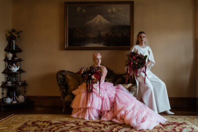 A couple sit on a vintage couch beneath a painting of Mt Hood. One of the partners wears a pink dress and the other wears a white dress. They both carry large floral bouqets