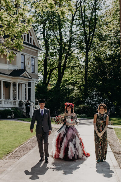 A bride in a colorful gown and hat is escorted to the ceremony by her parents. An ornate Victorian house stands in the background among tall trees.