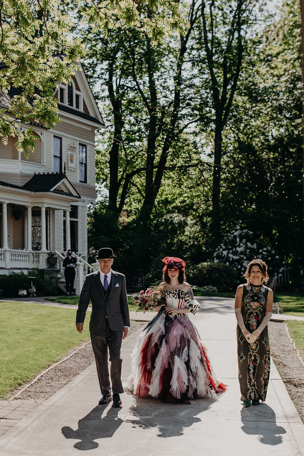 A bride in a colorful gown and hat is escorted to the ceremony by her parents. An ornate Victorian house stands in the background among tall trees.