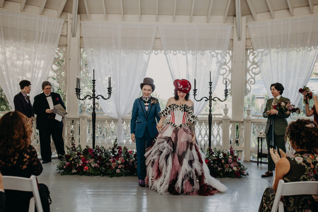 A bride in a blue suit and a bride in a colorful dress and red hat walk down the aisle after their wedding ceremony