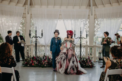 A bride in a blue suit and a bride in a colorful dress and red hat walk down the aisle after their wedding ceremony
