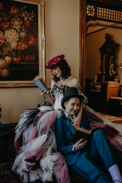 A bride in a multi-colored dress and a bride in a blue suit sit reading vintage books with a floral painting behind them.