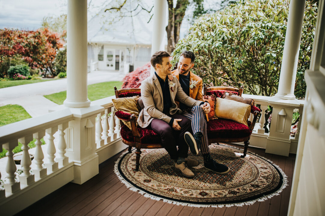 Two grooms sit on an antique velvet couch on the porch of a Victorian mansion. Behind them are shrubs, lawns, and a gazebo.