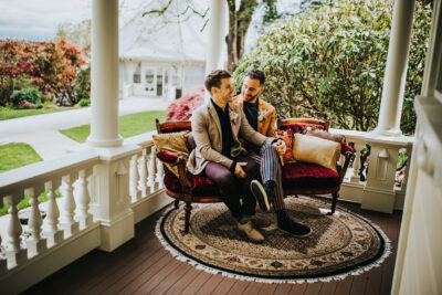 Two grooms sit on an antique velvet couch on the porch of a Victorian mansion. Behind them are shrubs, lawns, and a gazebo.