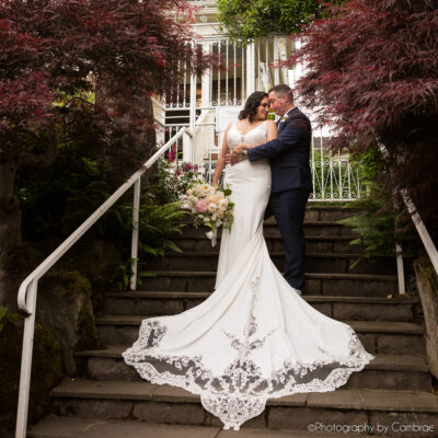 A bride and groom embrace on a stairway in front of a white iron gate. They are surrounded by maple trees.
