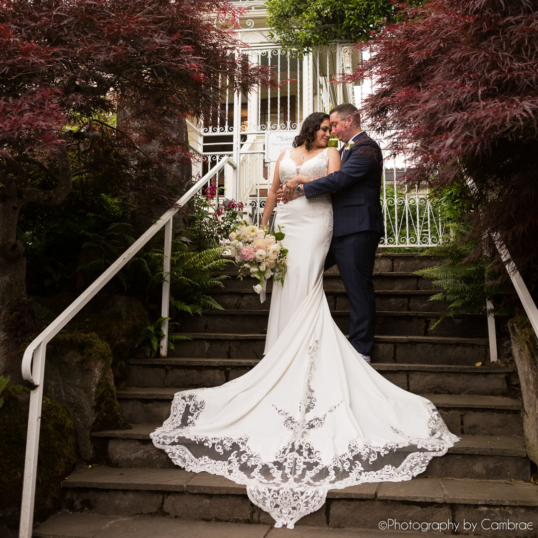 A bride and groom embrace on a stairway in front of a white iron gate. They are surrounded by maple trees.